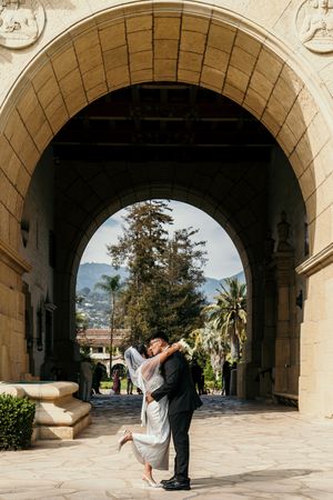 Bride and groom share a romantic dip kiss under a grand stone arch at an elegant outdoor wedding venue.