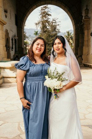 Bride in white gown and veil holds bouquet beside woman in blue dress under stone arch.