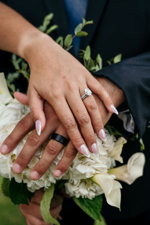 Newlyweds displaying wedding rings on hands resting over white hydrangea bridal bouquet.
