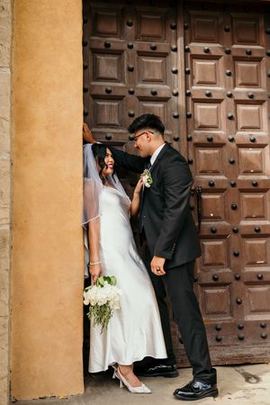 Bride and groom share a romantic moment by ornate wooden doors, bride holding white bouquet in elegant satin dress.