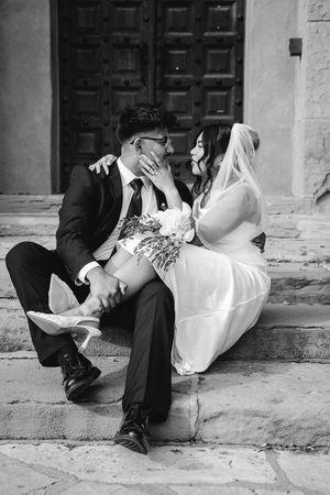 Black and white photo of a bride and groom sitting on stone steps, sharing an intimate moment with bridal bouquet.