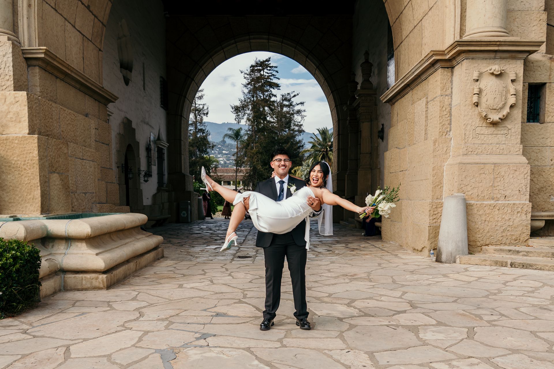Groom carries smiling bride through stone archway at elegant wedding venue with mountains behind.