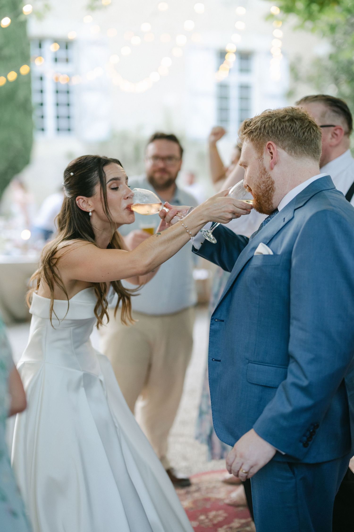 Bride laughing joyfully holding champagne glass while dancing with guests at outdoor wedding reception.