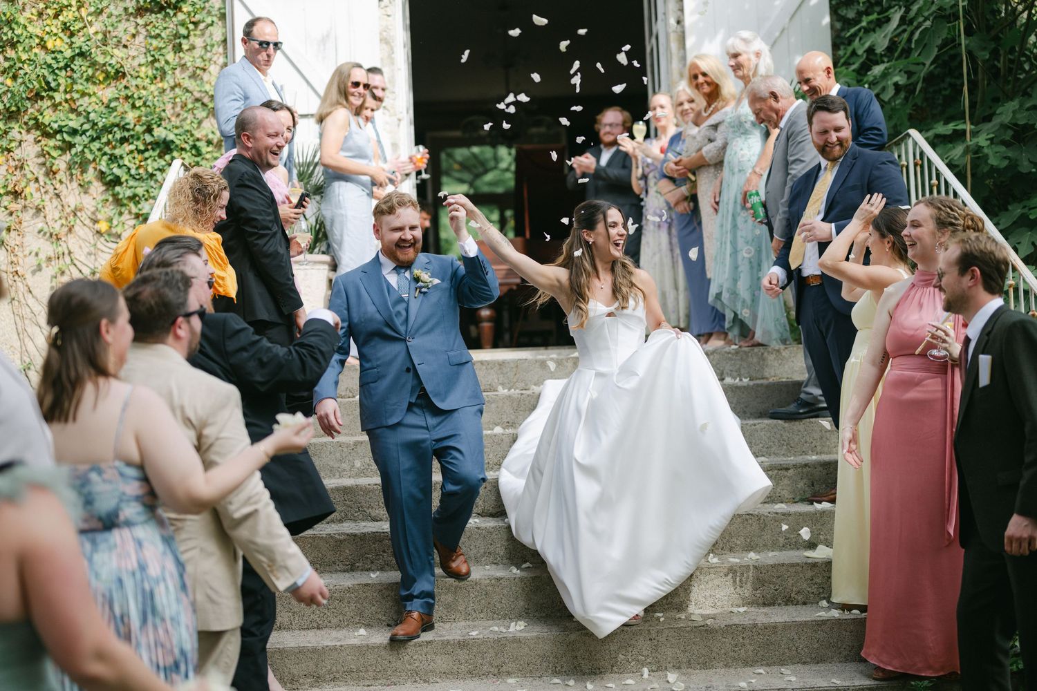 Bride and groom walk down stone steps as guests throw confetti at their outdoor wedding celebration.