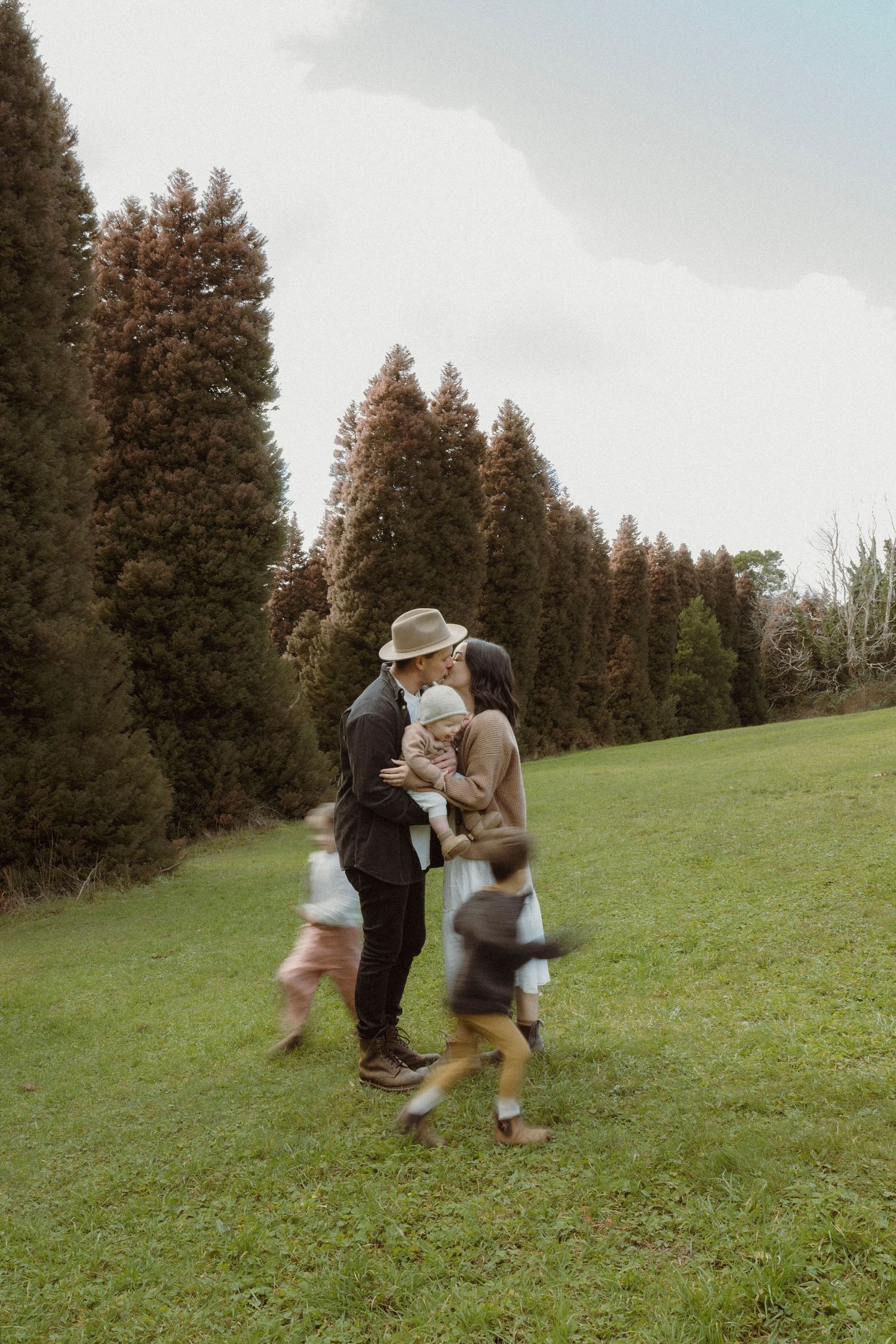 A couple shares a tender moment holding a baby on a green lawn, while two children run around them near tall evergreen trees.