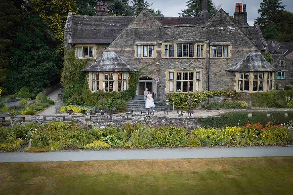 Bride and groom pose in front of a grand stone manor house with lush gardens in a scenic countryside setting.
