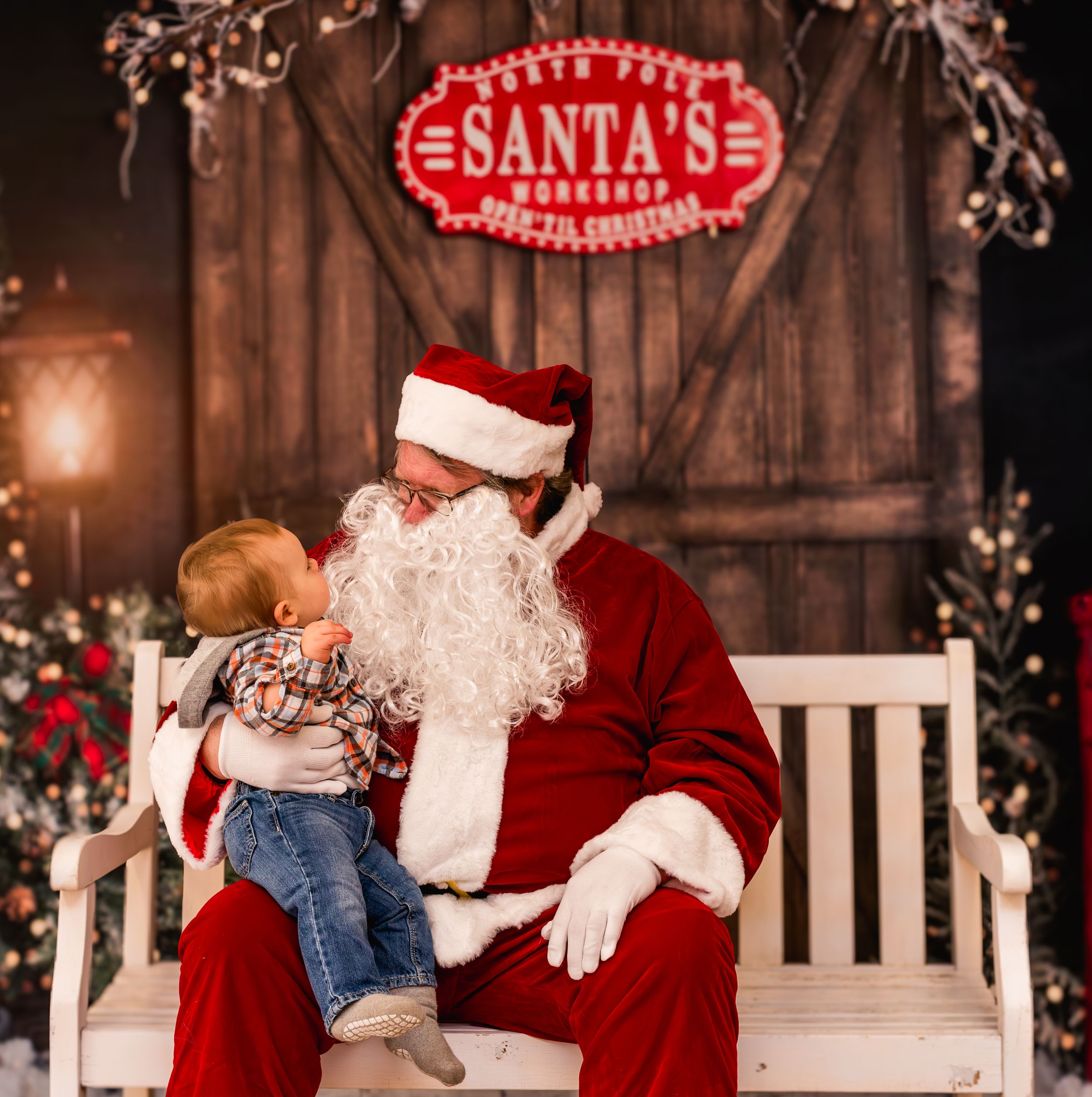 A toddler sits on Santa Claus's lap in front of a festive North Pole workshop backdrop during a Christmas photo session.