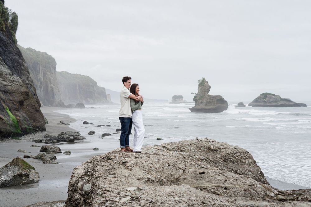 Couple standing hugging looking out to sea with the rugged West Coast landscape, north of greymouth