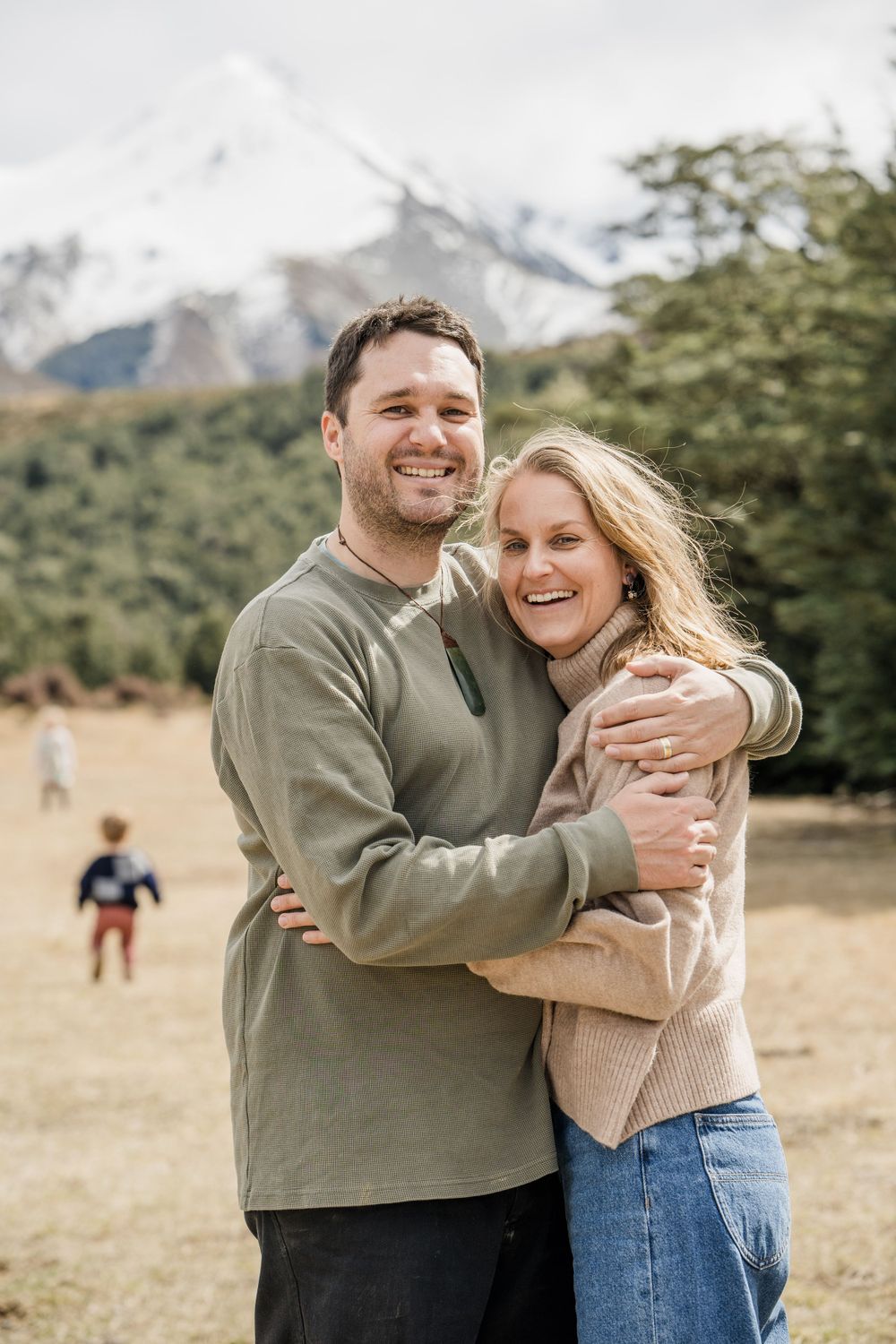 Couple standing cuddling and smiling at the camera with children running in the background, at Castle Hill