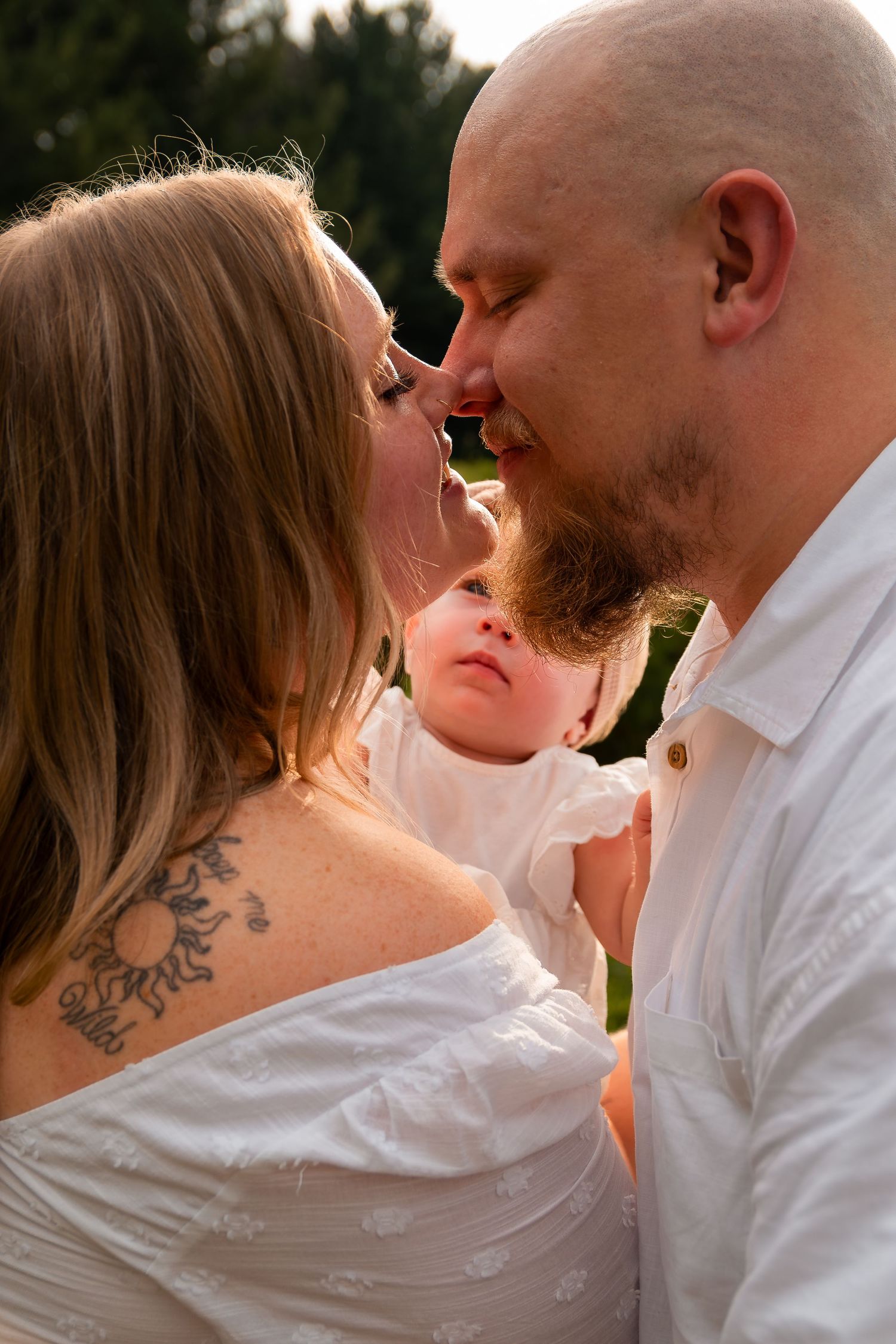 A couple in white shares a tender nose kiss while holding their newborn baby outdoors in soft golden light.
