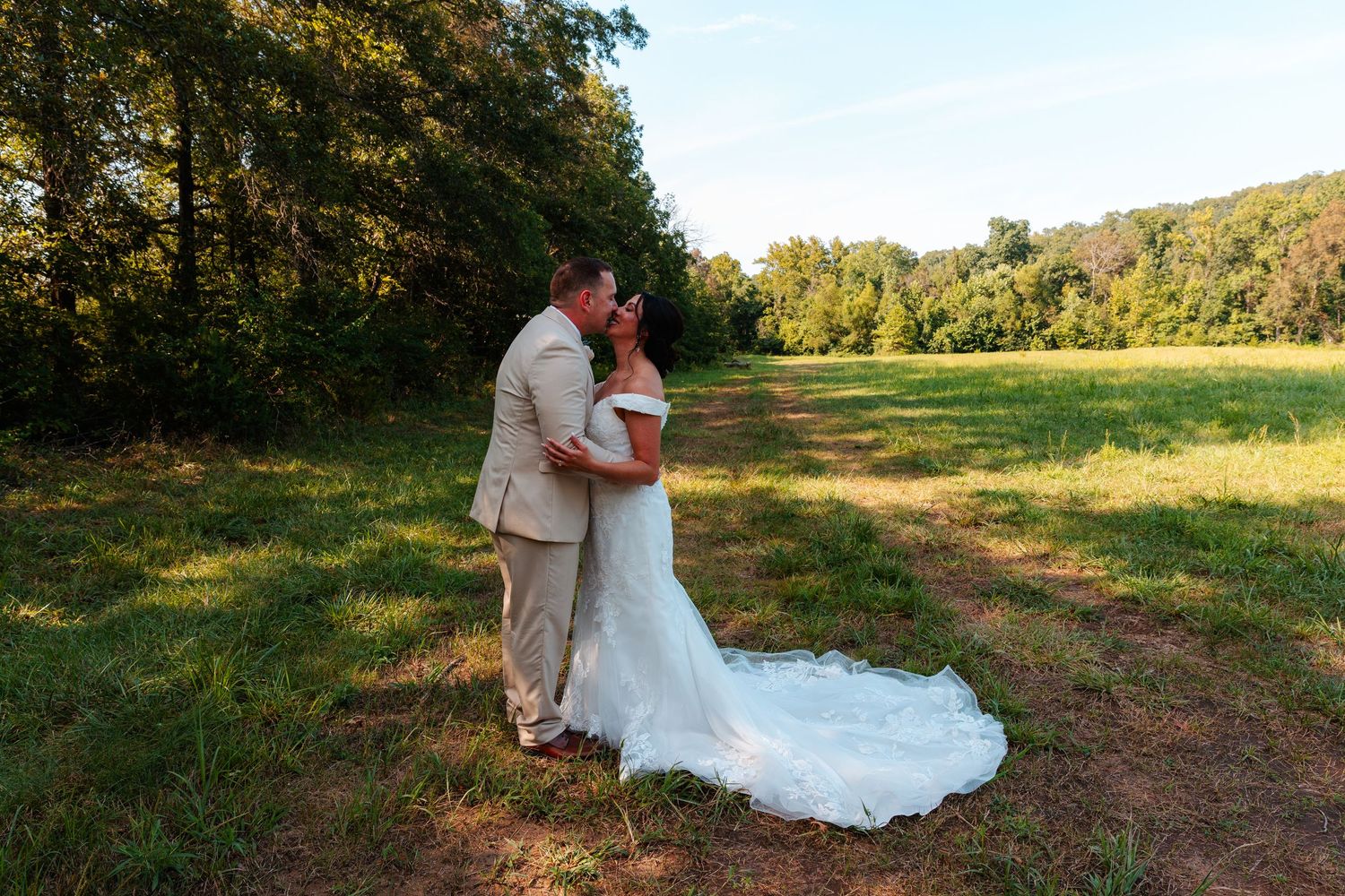 Bride and groom share a romantic kiss in a sunlit green meadow surrounded by lush trees on their wedding day.