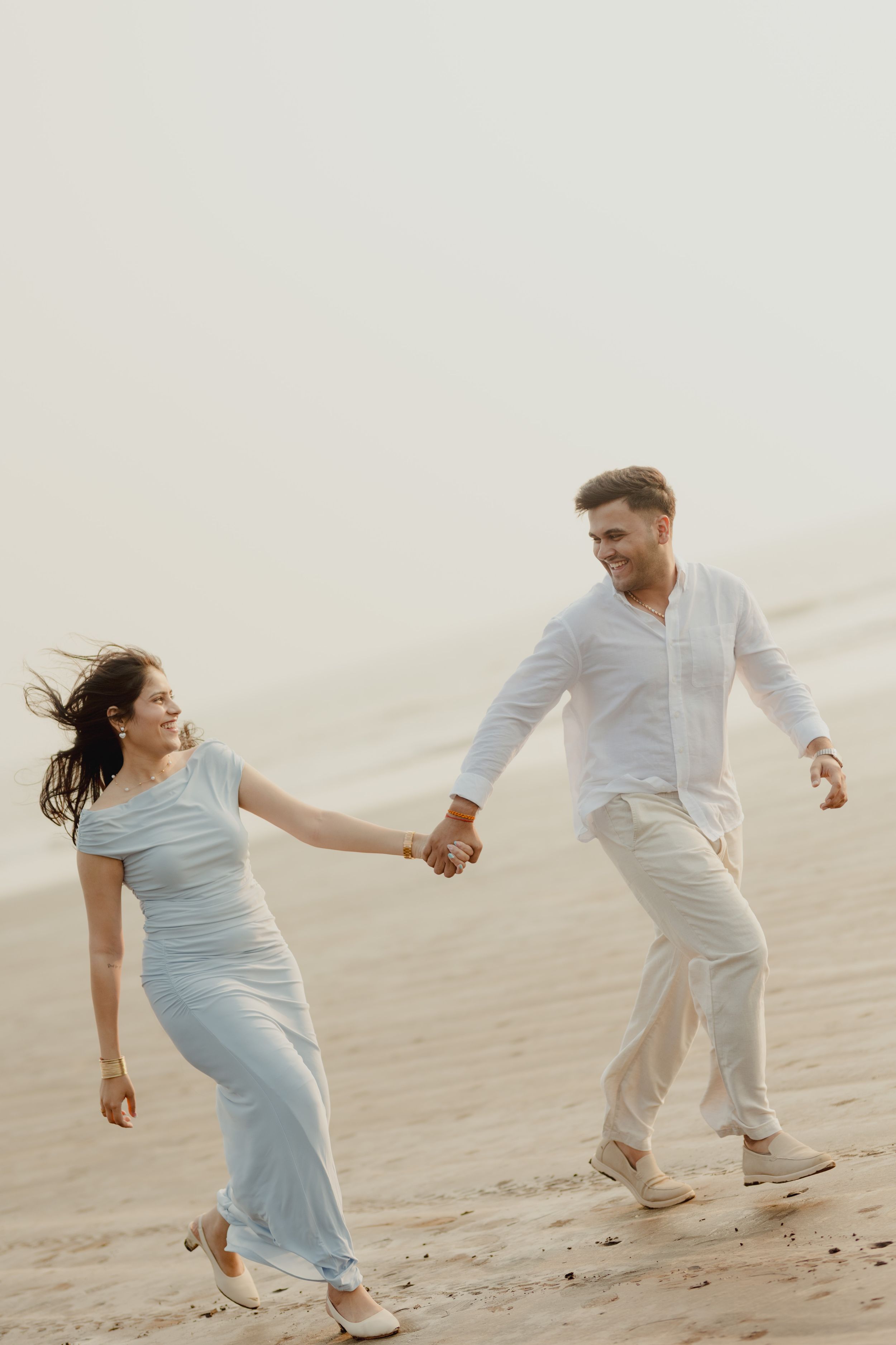 A couple holds hands and walks joyfully across sandy dunes, woman in light blue dress, man in white outfit.