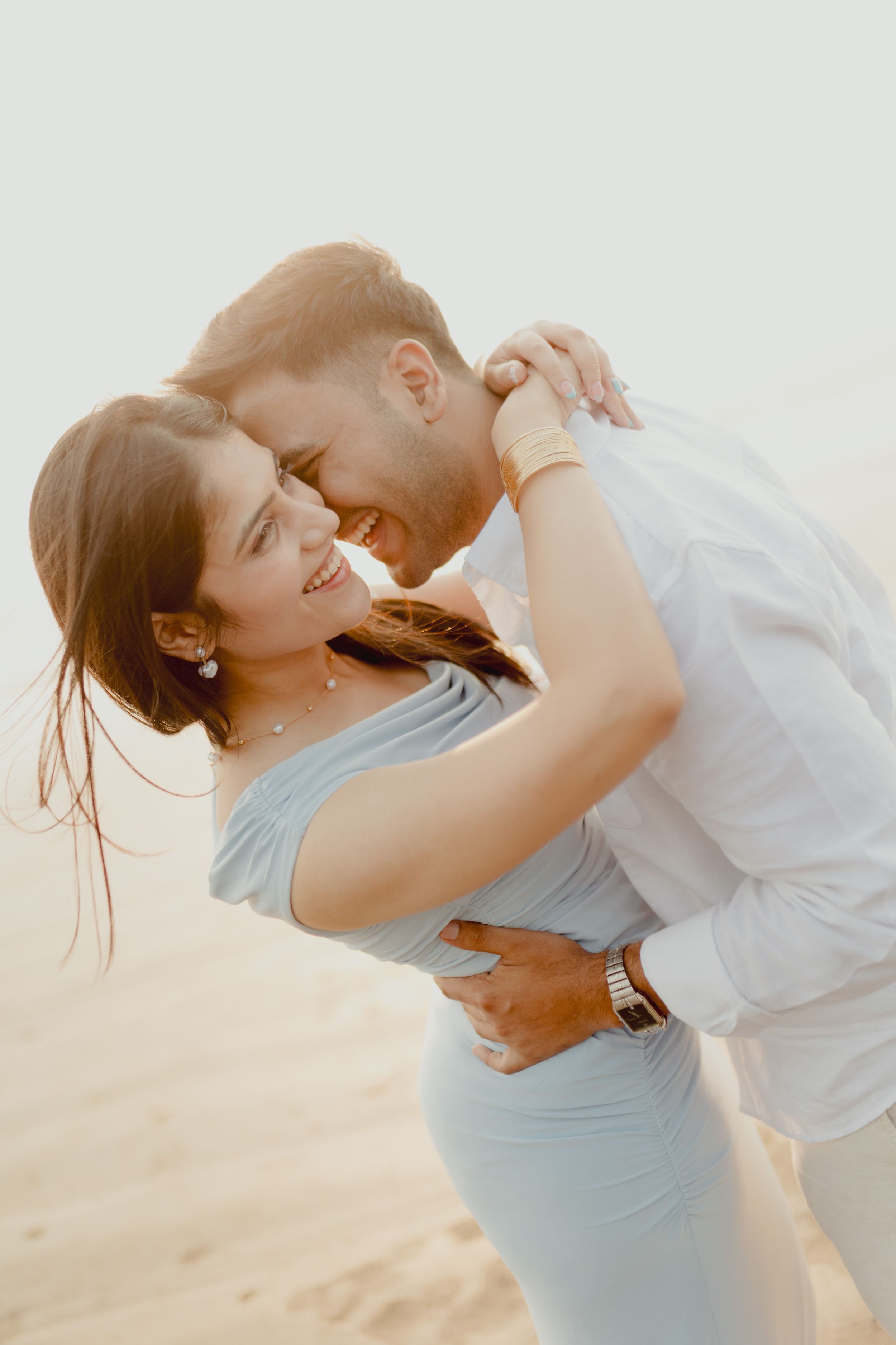A joyful couple embraces and smiles on a sandy beach, wearing light blue and white outfits in soft natural light.