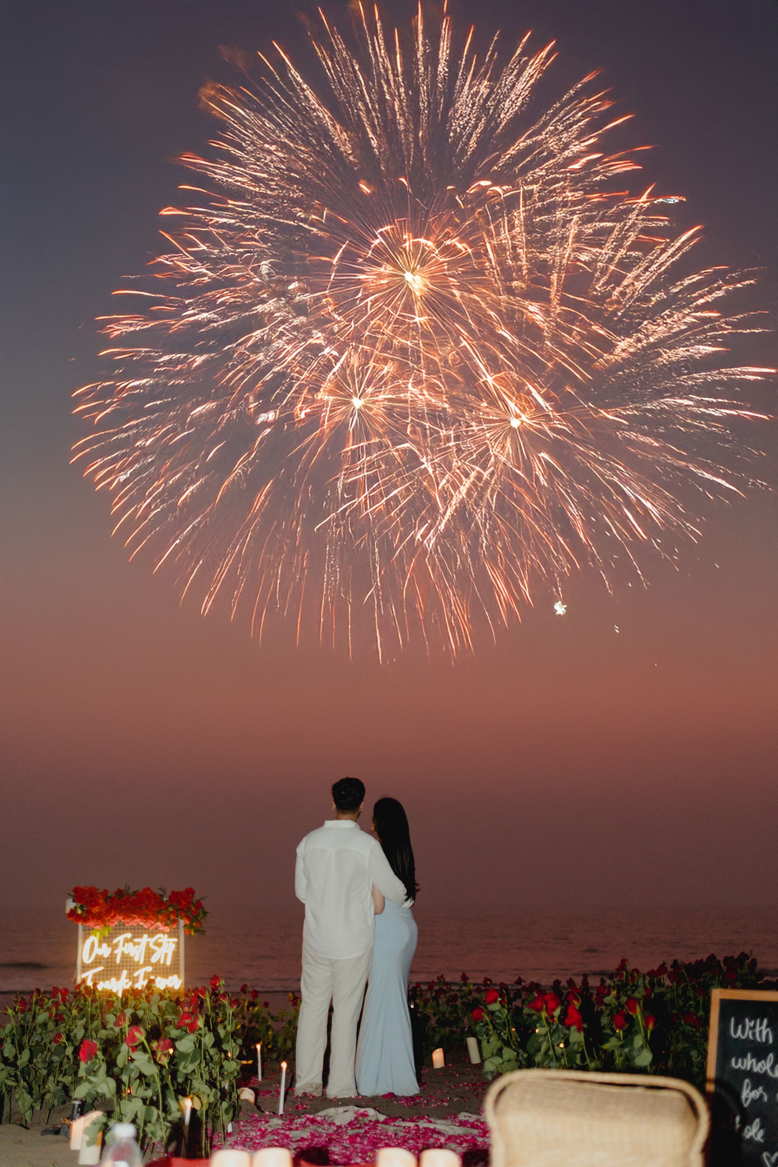 Couple watches stunning fireworks display on a romantic rose-filled beach with neon sign at sunset.