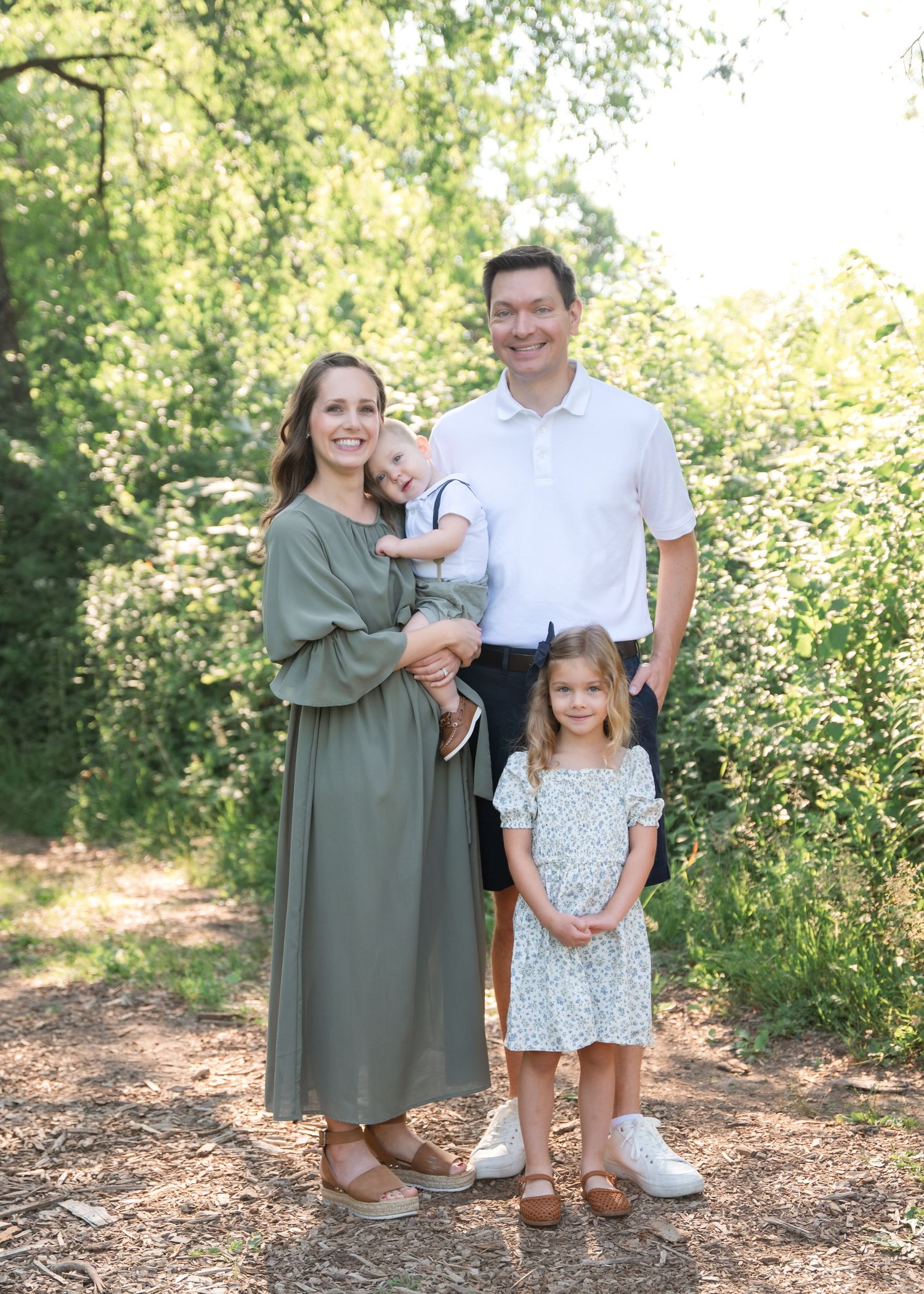 Family of four posing on a wooded path, mother in sage dress holding toddler, father in white polo, young girl in floral dress.