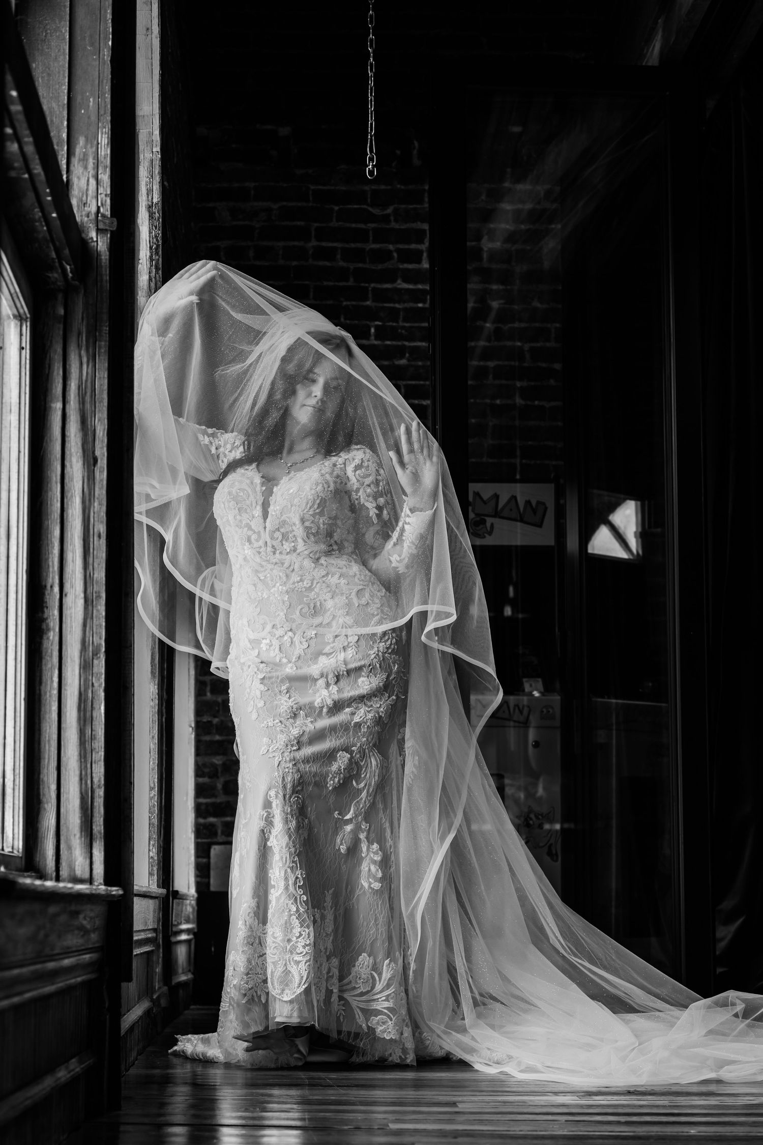 Black and white photo of a bride in a lace gown with veil flowing overhead in an elegant indoor setting.