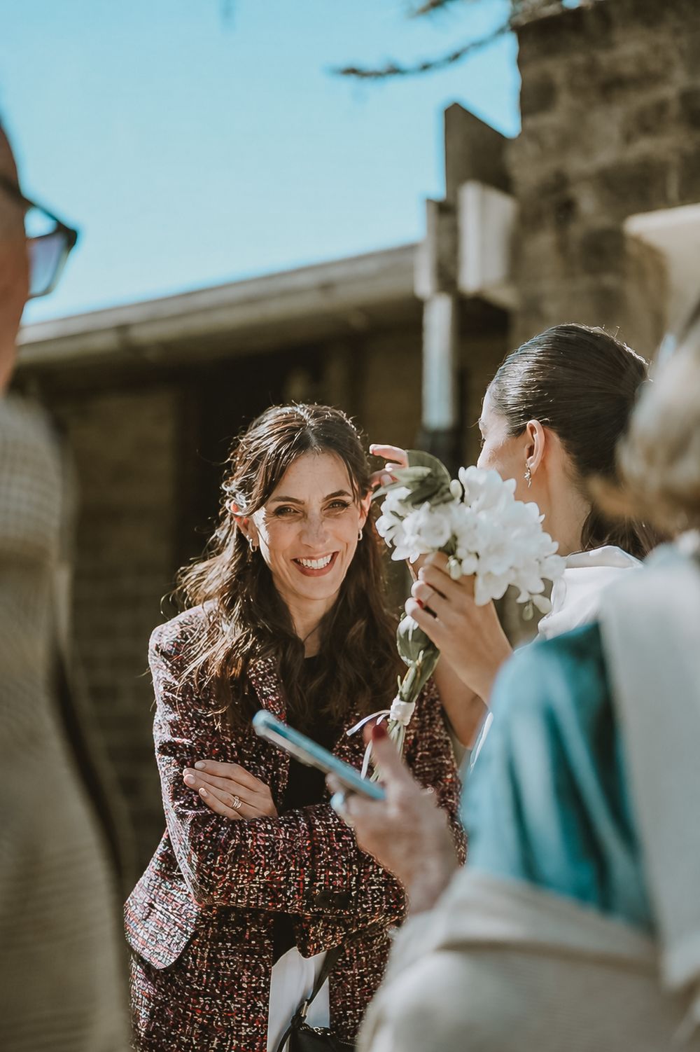 Smiling woman in floral jacket greeting the bride holding white flowers at outdoor wedding.