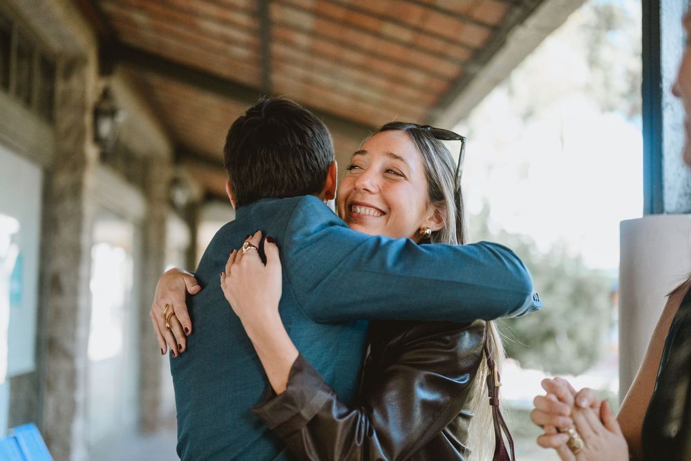 A woman smiling joyfully while hugging someone in a teal shirt outdoors near a rustic building.