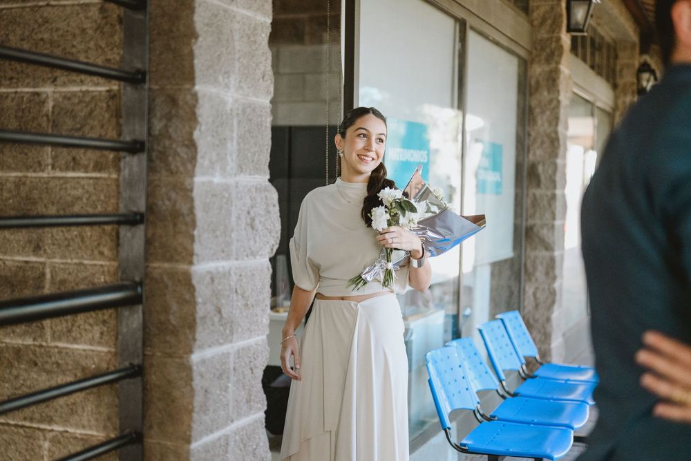 Bride smiling holding white flower bouquet in cream dress at venue entrance.