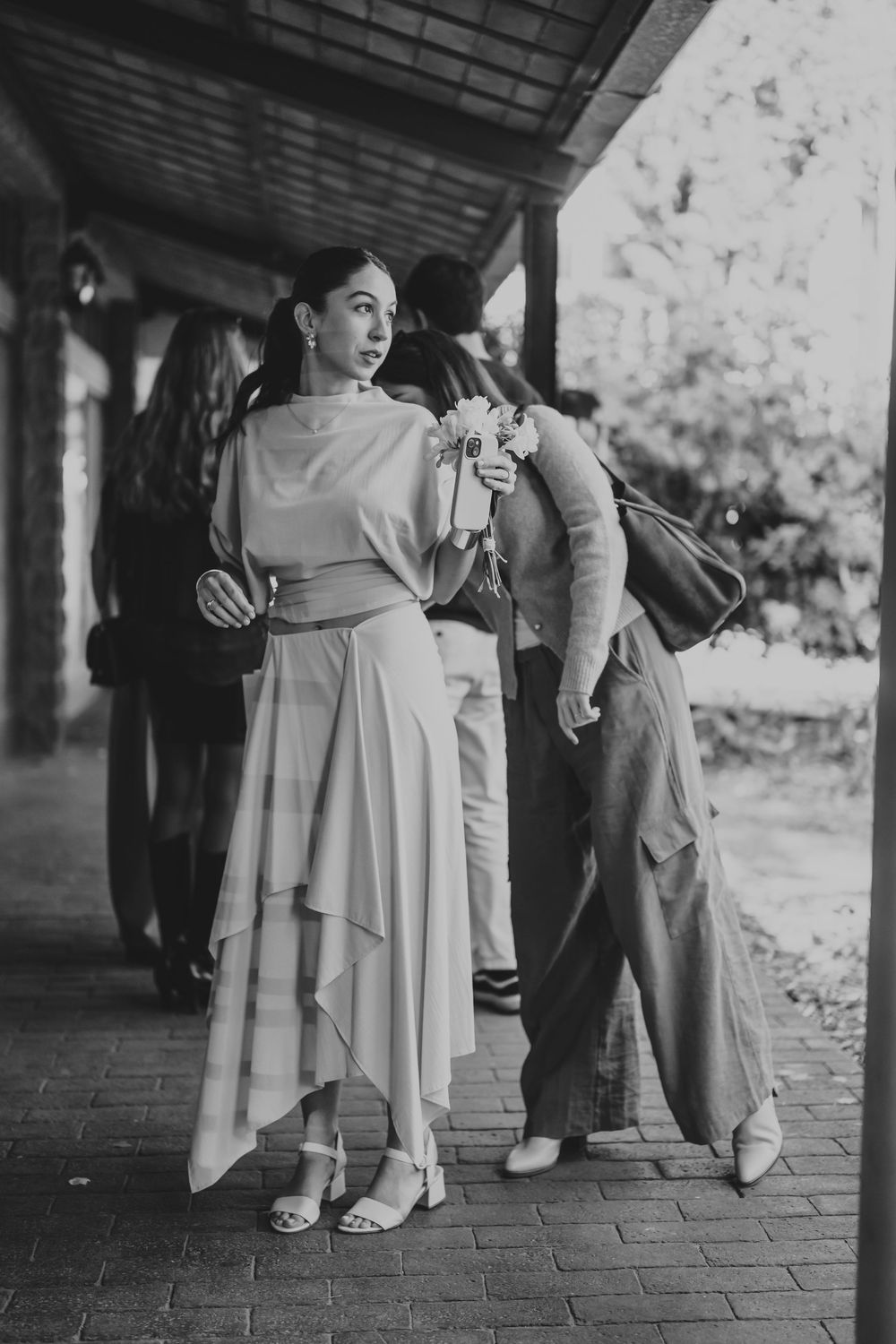 Elegant bridesmaid in flowing dress holds bouquet while standing under covered walkway in black and white photo.