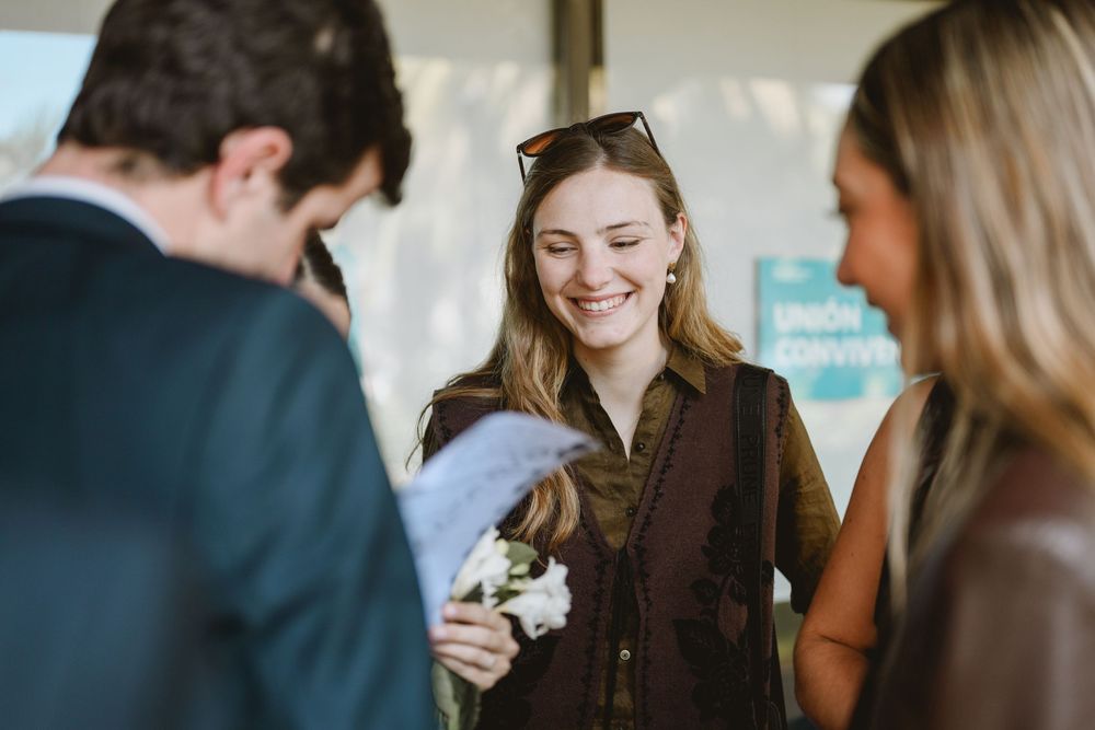 Smiling blonde woman holds white flowers while chatting with guests at an outdoor wedding event.