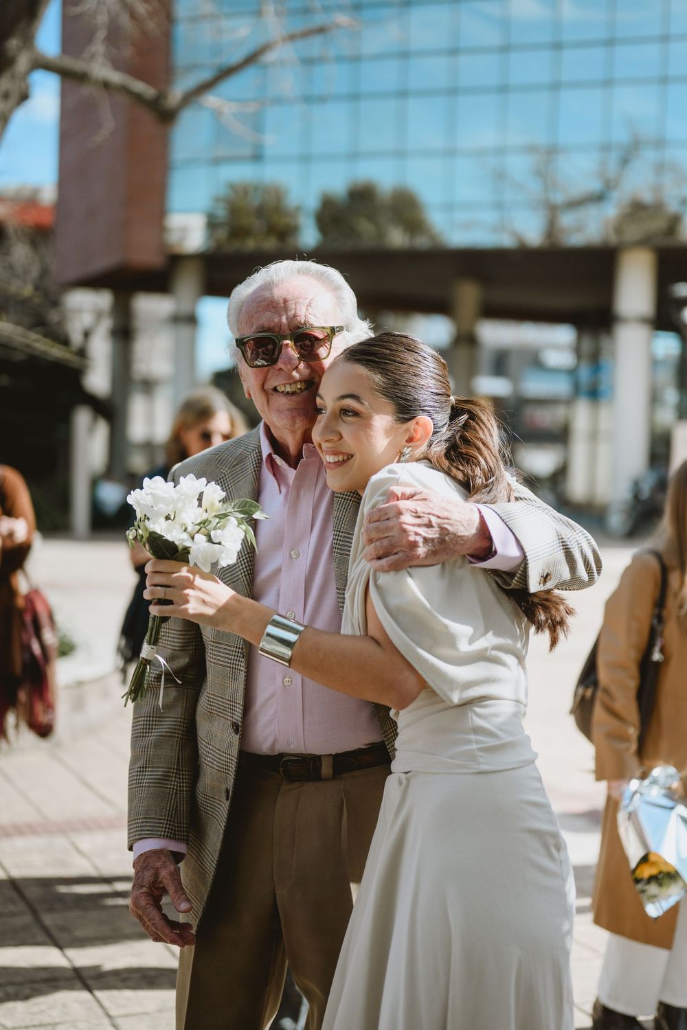 Elderly man in suit hugs smiling young woman in white dress holding white flowers outdoors on sunny day.