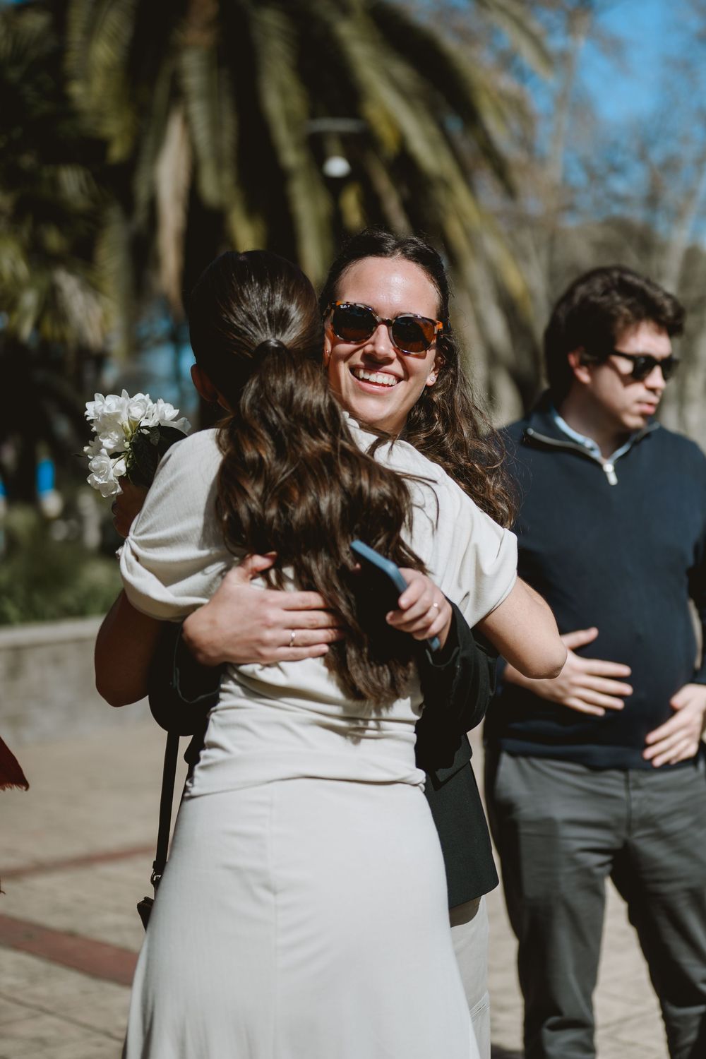 Smiling bride in white dress hugging a guest outdoors while holding white flowers at wedding.