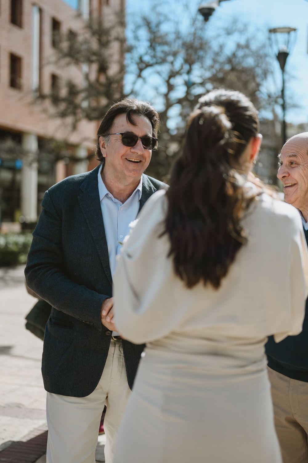 Man in dark blazer and sunglasses smiling warmly while greeting guests outdoors on a sunny day.