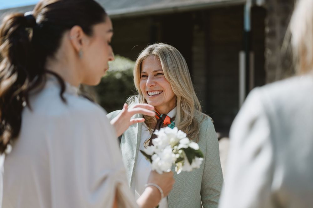 Smiling blonde woman holding white flowers while chatting with guests at an outdoor event.