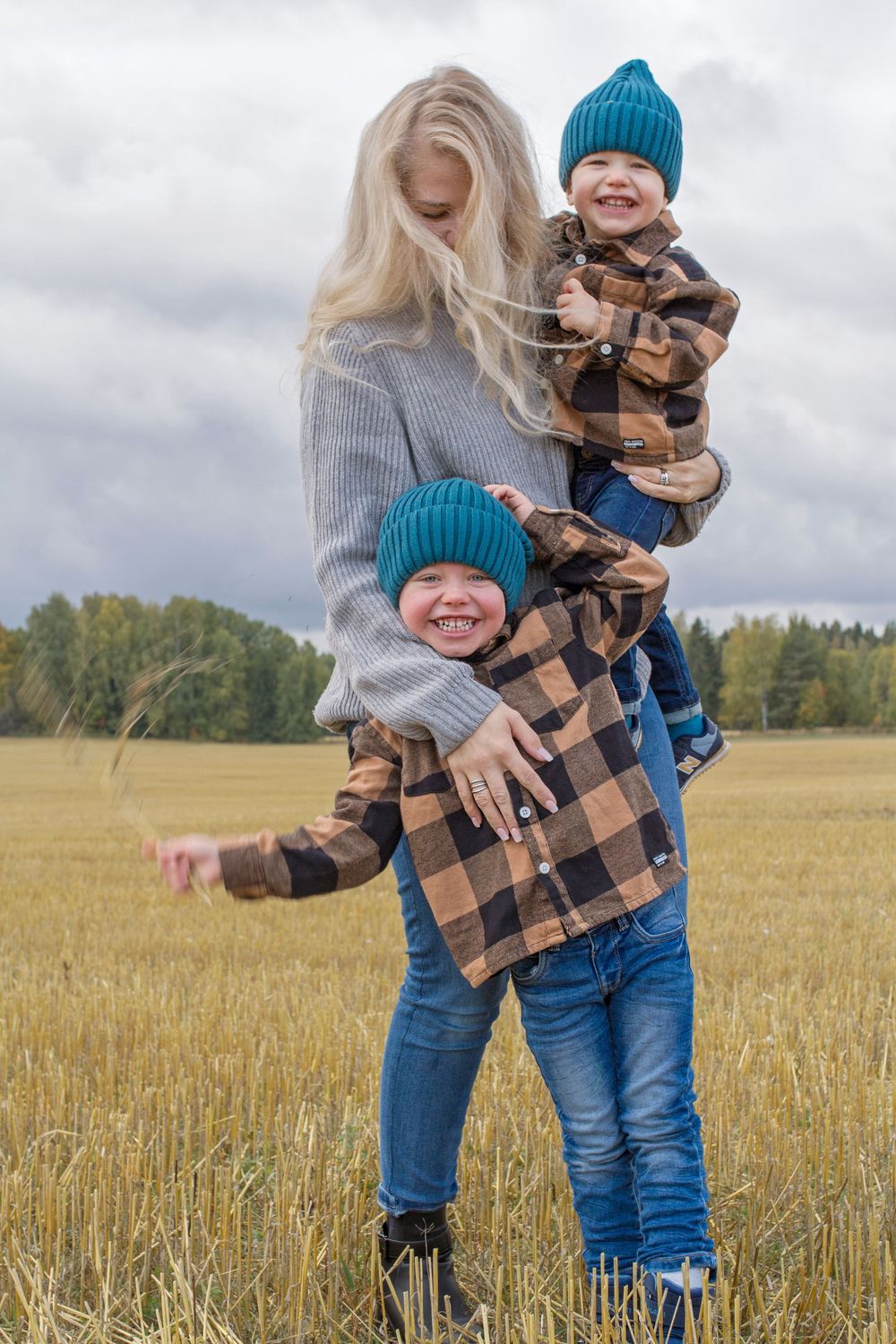 A mother holds two smiling boys in matching plaid shirts and teal hats in an autumn field.