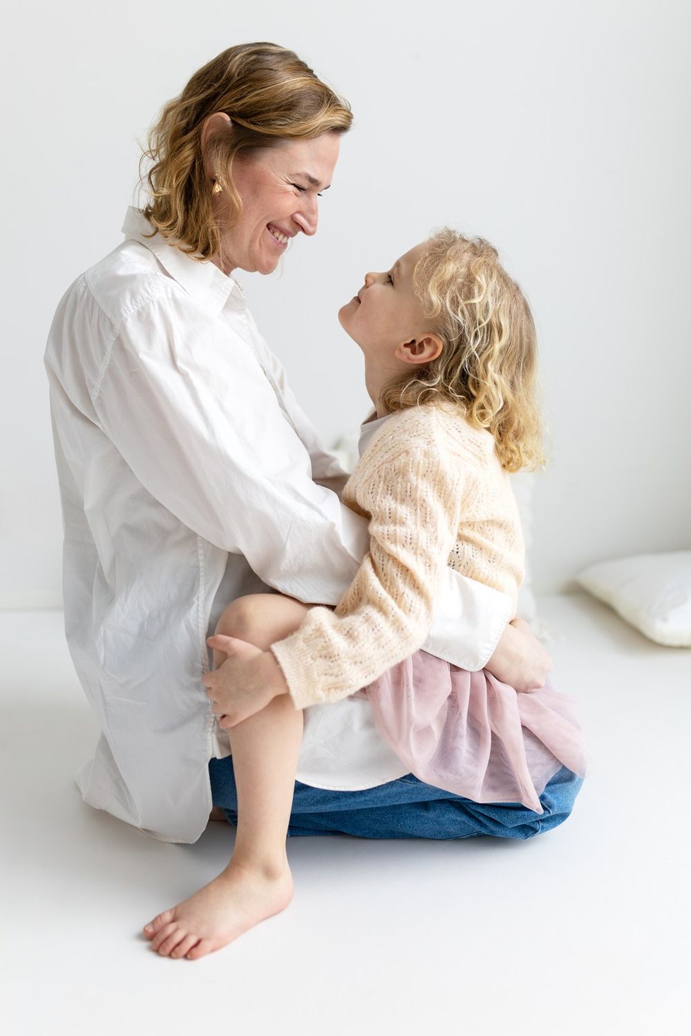 A smiling mother sits on the floor hugging her laughing toddler daughter, both wearing white tops in a bright room.