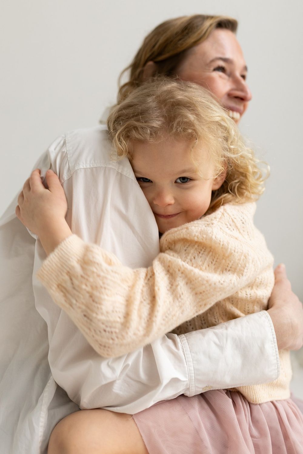 A young curly-haired girl in a pink sweater hugs a smiling woman in a white shirt against a light background.