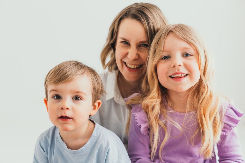 A smiling woman poses with two young children, a boy and a girl with blonde hair, against a white background.