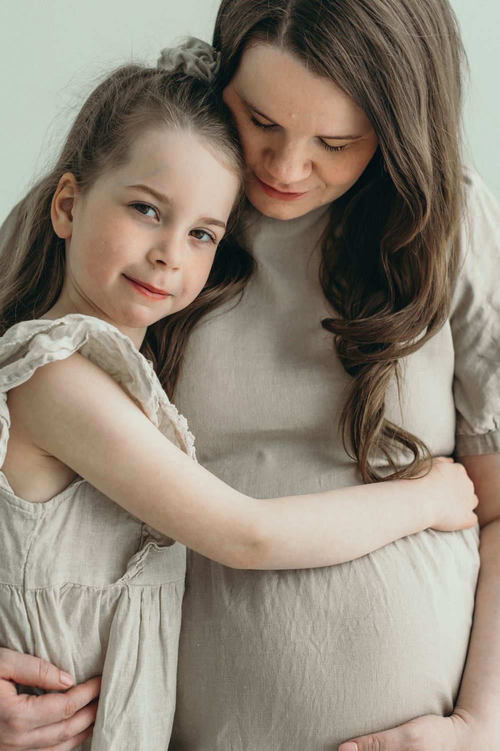 A young girl hugs her pregnant mother tenderly, both wearing matching beige linen dresses in a soft studio setting.
