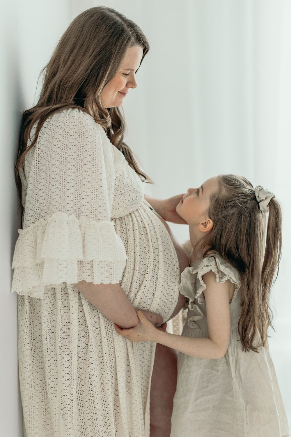 Pregnant woman in lace dress smiling at young daughter who touches her baby bump in soft white studio.