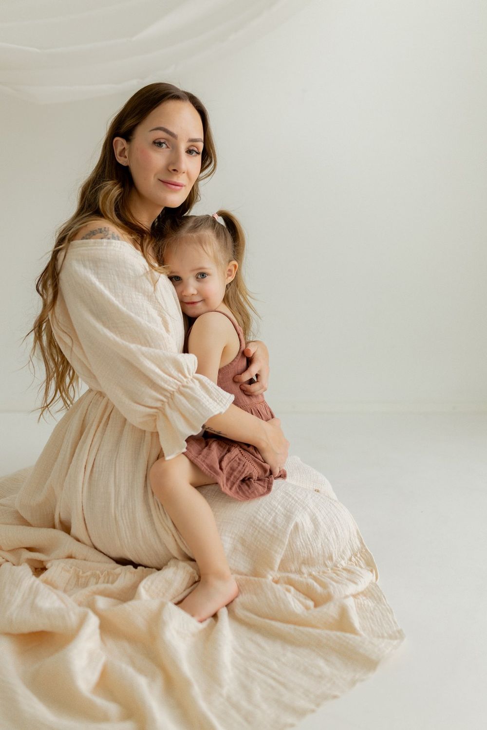 A mother in a cream dress tenderly holds her young daughter in a soft, light-filled studio setting.