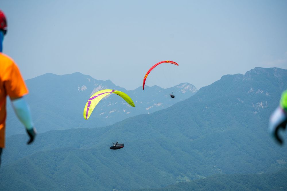 Colorful paragliders soaring over lush green mountains on a clear day, with spectators watching from the foreground.