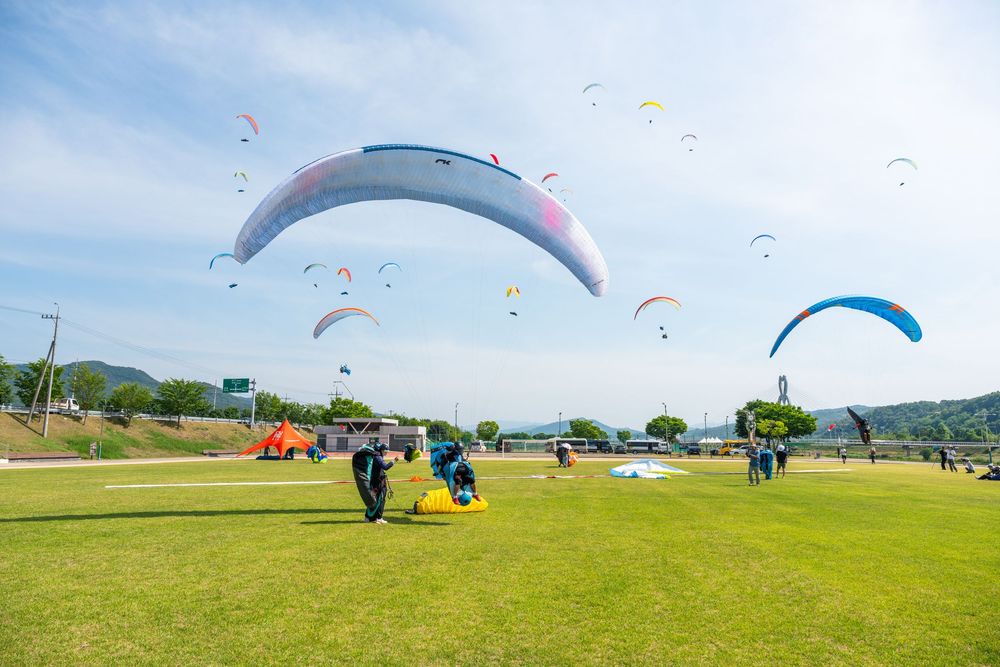Paragliders launching colorful canopies on a green field under a blue sky with multiple gliders soaring above.