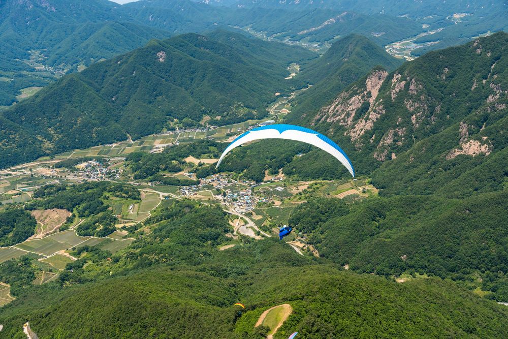 Paraglider soaring over a lush green valley with a small village and mountain ridges in South Korea.