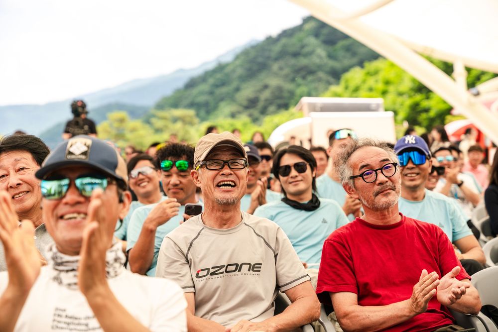 Crowd of smiling men clapping outdoors at an event, with mountains and trees in the background.
