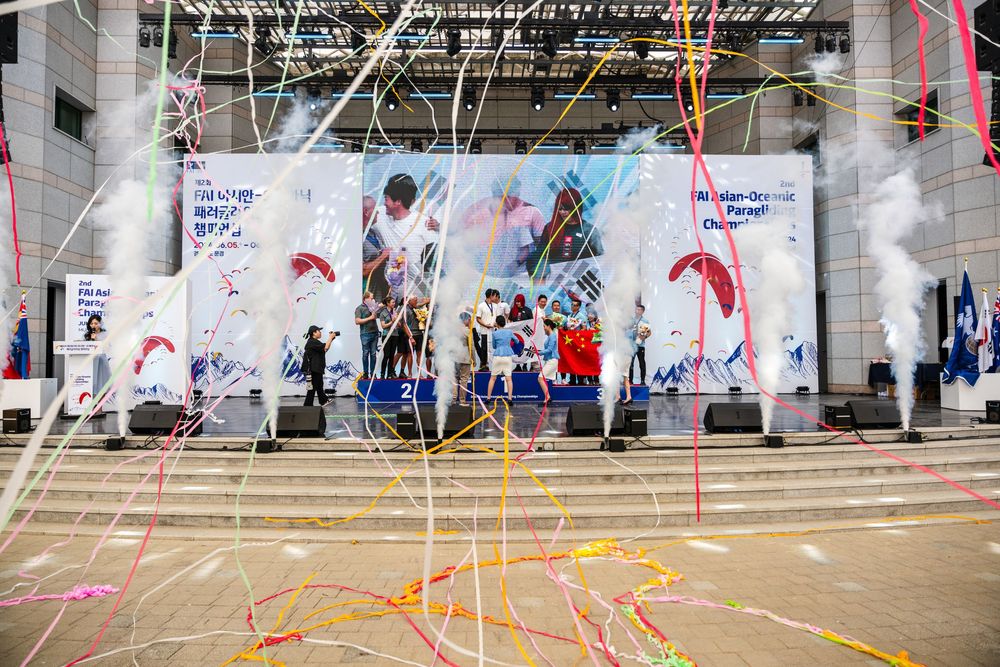 Colorful streamers and confetti fill the air at a festive outdoor stage event with performers and a vibrant backdrop.