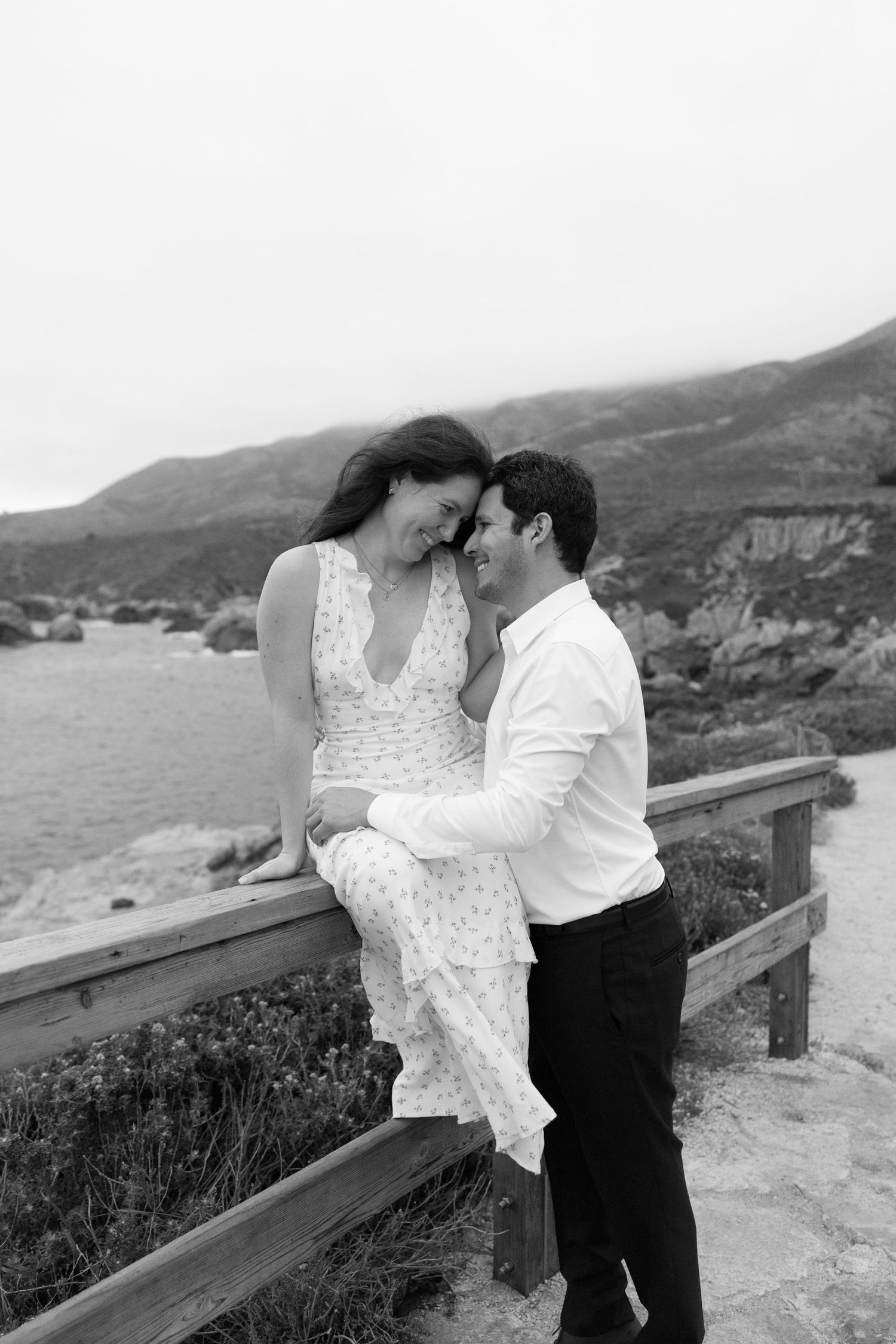 Black and white photo of a romantic couple embracing on a coastal fence with scenic mountains and ocean in the background.