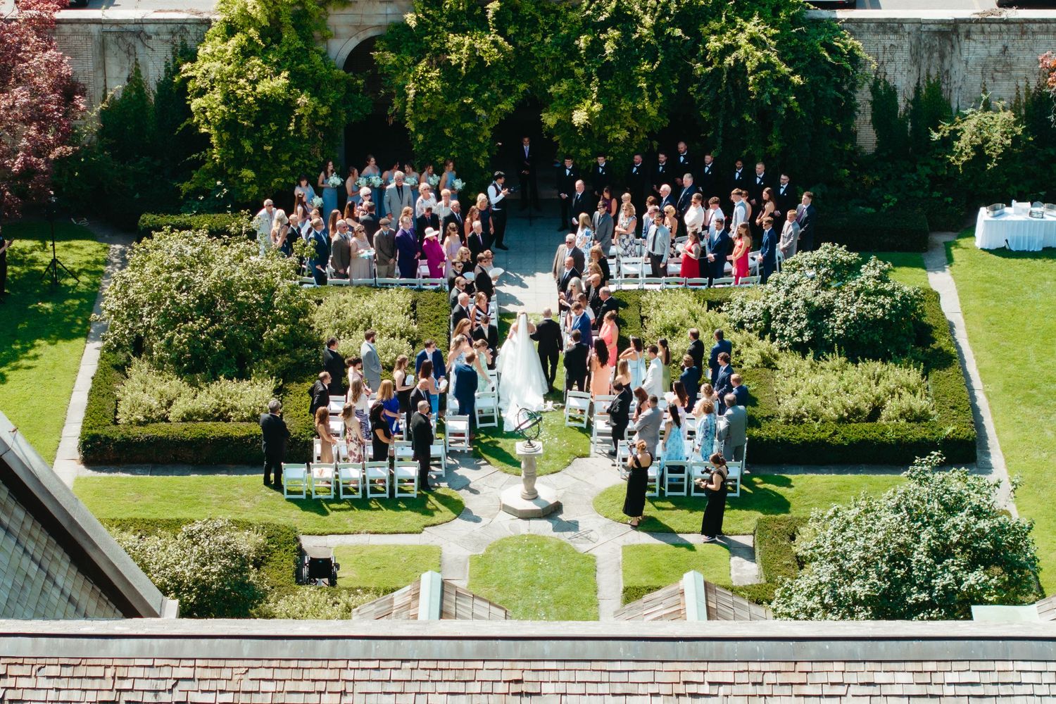 Aerial view of an elegant outdoor wedding ceremony in a lush garden courtyard with guests seated in rows.