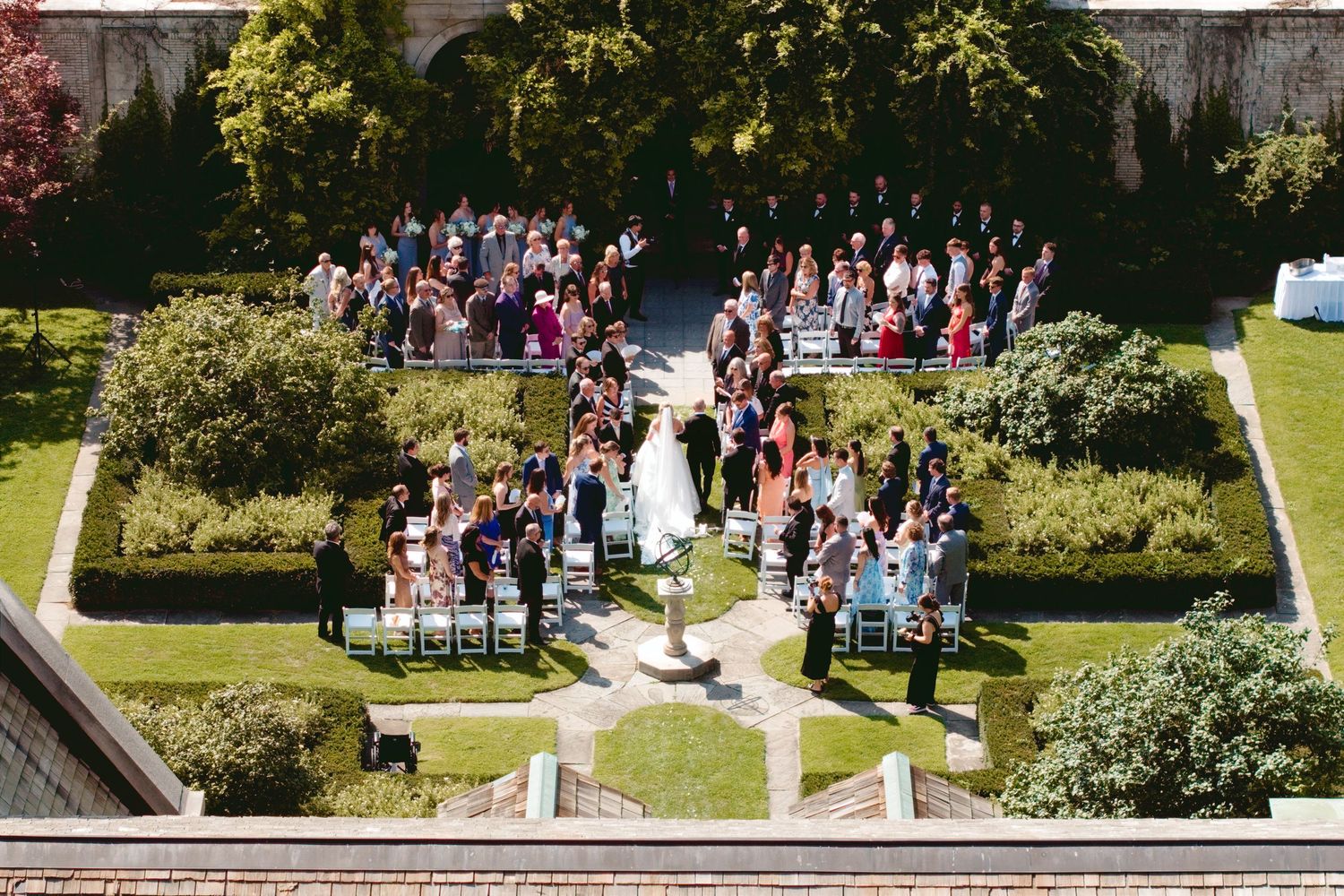 Aerial view of an elegant outdoor wedding ceremony in a lush garden courtyard with guests seated in rows.
