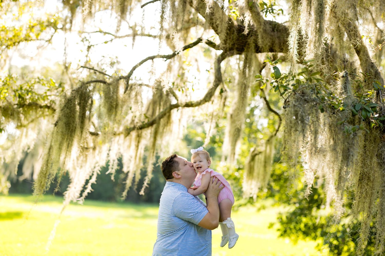 Father kissing baby under Spanish moss-draped oak tree in sunny park setting.