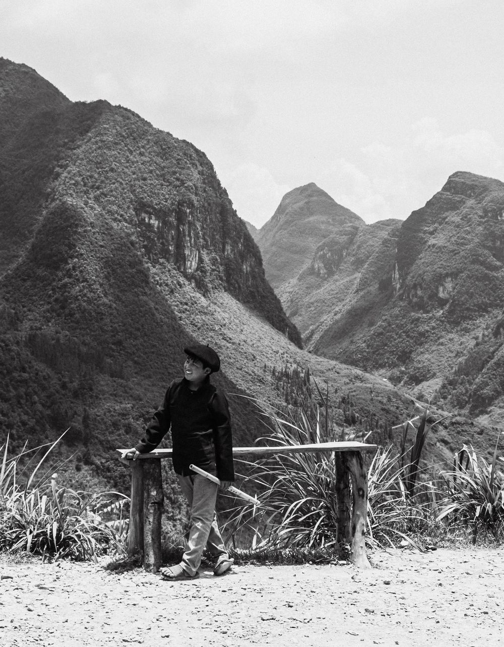 Black and white photo of a person leaning on wooden posts with dramatic mountain valley backdrop.