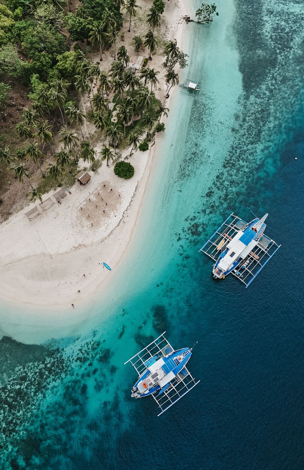 Aerial view of tropical island beach with turquoise water and traditional outrigger boats anchored offshore.