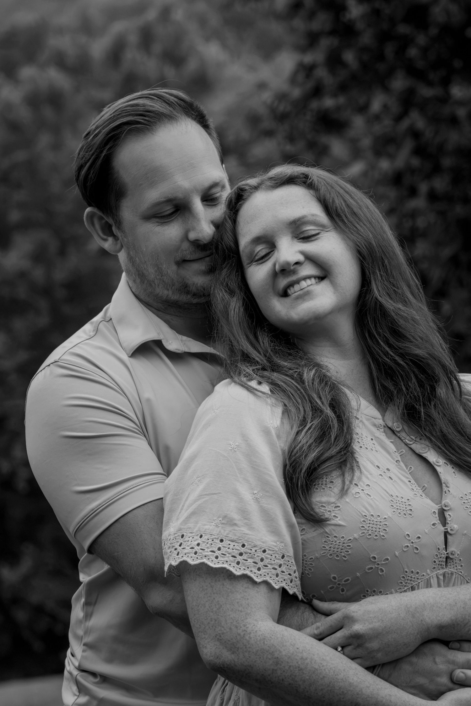Black and white photo of a couple embracing outdoors, woman smiling with eyes closed as man nuzzles her cheek.