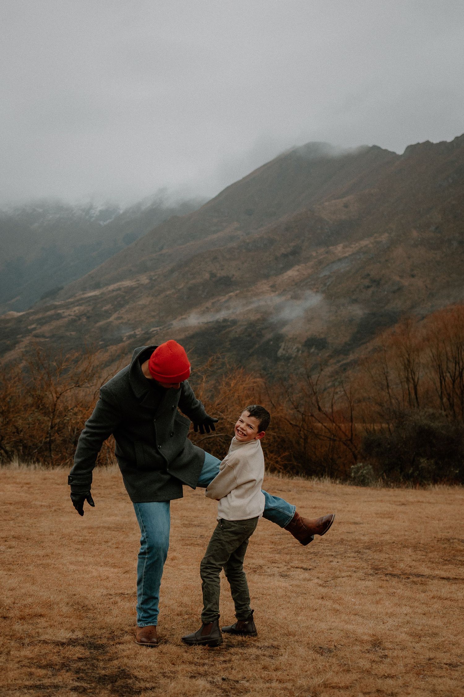 A parent and child playfully dancing in a misty mountain landscape during autumn, wearing warm winter clothing.