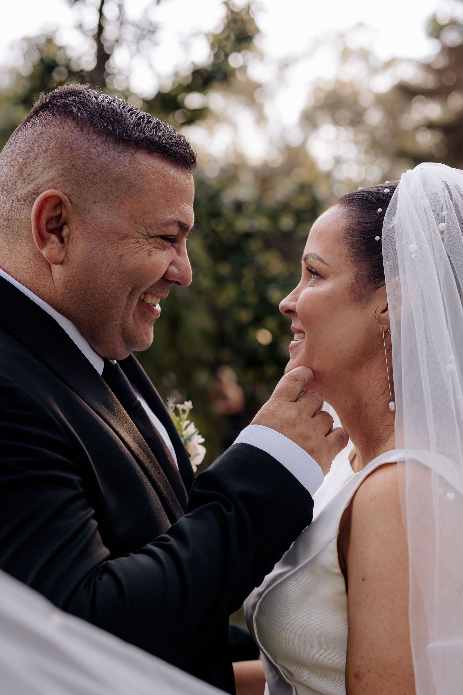 Smiling bride and groom share a tender moment, groom gently touching bride's chin during outdoor wedding ceremony.